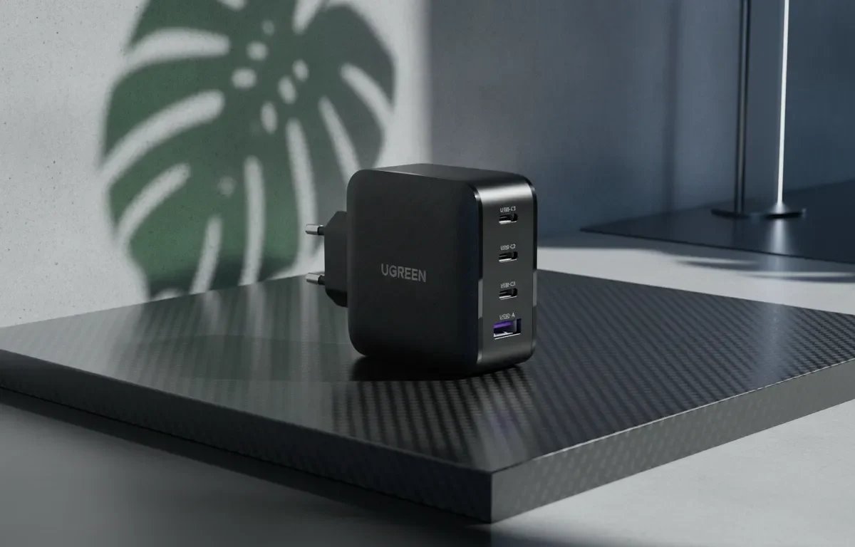 Carregador preto UGREEN de 4 portas sobre base de fibra de carbono com sombra de folha ao fundo.