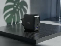 Carregador preto UGREEN de 4 portas sobre base de fibra de carbono com sombra de folha ao fundo.