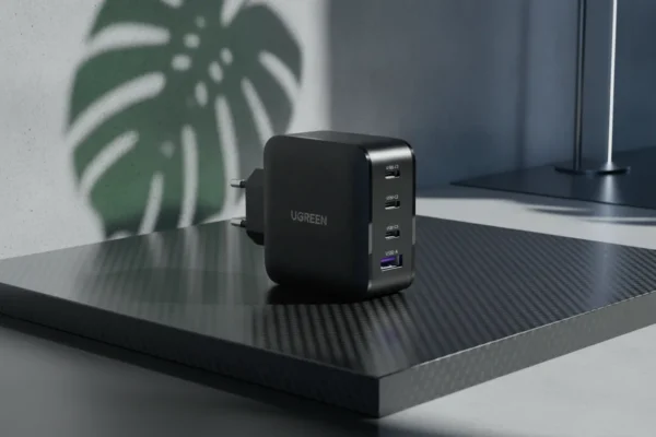 Carregador preto UGREEN de 4 portas sobre base de fibra de carbono com sombra de folha ao fundo.