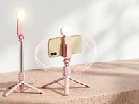 Tripé bastão de selfie rosa com celular posicionado na horizontal e mini ring light acesa em um cenário de estúdio moderno desfocado.