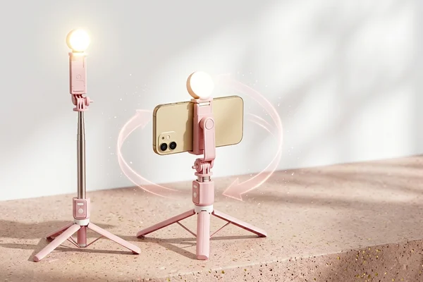 Tripé bastão de selfie rosa com celular posicionado na horizontal e mini ring light acesa em um cenário de estúdio moderno desfocado.