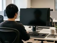 Pessoa de costas diante de um monitor apagado sobre uma mesa de trabalho, em um ambiente com luz natural