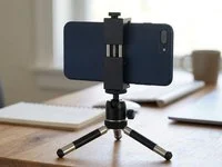 Celular de tela apagada preso a um suporte metálico sobre um tripé de mesa, em ambiente iluminado